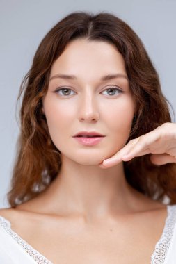 Işıl ışıl cildi ve sakin ifadesi olan genç bir kadın, elleri çenesinin altında, yumuşak beyaz bir elbise giyerek poz veriyor. Minimalist güzellik reklamları, cilt bakımı markaları ve sağlık başyazıları için ideal..