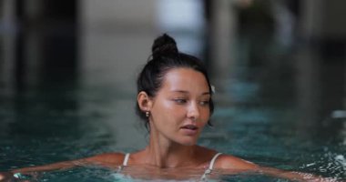 Beyaz mayo giymiş bronzlaşmış bir kadın havuzda nazikçe süzülüyor, saçlarında çiçek, sakin ve yavaş çekimde çekiliyor. Güzellik, spa, rahatlama, sağlık ve lüks yaşam tarzı içeriği için mükemmel..