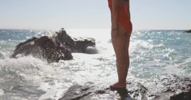 Kırmızı mayo giymiş manken, etrafındaki okyanus dalgaları deniz kenarındaki bir kayanın üzerinde güçlü bir şekilde duruyor. Güzellik, güven, seyahat, spor ve mayo kampanyaları için yavaş çekim ideal..