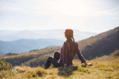 Kırmızı şapkalı ve ekose gömlekli bir kadın yürüyüşçü çimenli bir tepede oturur ve Karpat Dağları 'nın nefes kesici manzarasını seyreder. Macera, rahatlama ve doğa kavramları için idealdir..