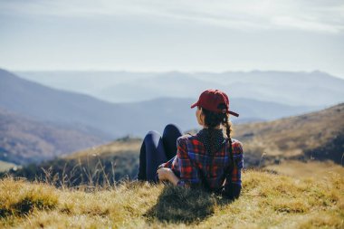 Kırmızı şapkalı ve ekose gömlekli bir kadın yürüyüşçü çimlerin üzerinde dinleniyor ve panoramik dağ manzarasının tadını çıkarıyor. Bu yüksek kaliteli görüntü seyahat, rahatlama ve doğa kavramları için mükemmeldir.