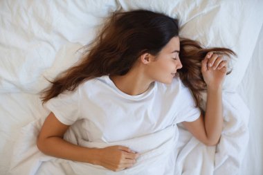 Huzurlu genç bir kadın beyaz çarşaflı bir yatakta mışıl mışıl uyur. Sabah güneşiyle yakalanır. Huzurlu ifadesi ve rahat ayarları dinlenme, rahatlama ve rahatlama temaları için ideal.