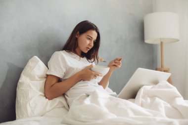 Genç bir kadın, sıcak ışıkla aydınlatılmış laptopuyla yatakta çalışırken kahvaltıdan zevk alıyor. Konforlu ortam, uzak çalışma, rahatlama ve modern yaşam tarzının temaları için mükemmel.