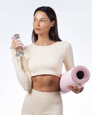 Elinde pembe yoga minderi ve su şişesi tutan bej renkli aktif giysili beyaz bir kadın başka tarafa bakıyor. Formunu, sağlığını, su ihtiyacını ve sağlıklı yaşam tarzını desteklemek için ideal. Temiz stüdyo geçmişi.