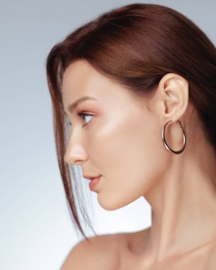 Yandan görünüşü kusursuz, parlak tenli, parlak kahverengi saçlı, geniş halka küpeli, cilt bakımı, mücevher, moda ya da güzellik ürünleri reklamı için mükemmel genç beyaz bir kadın..