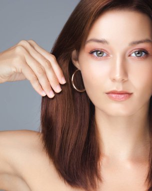 Kendine güvenen kızıl saçlı, parlak saçlı ve halka küpeli bir kadın saçına hafifçe dokunuyor. Saç bakımı, saç boyama ya da güzellik kampanyaları için mükemmel. Temiz stüdyo geçmişi ve modern tarzı.