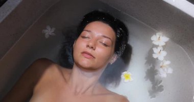 Beyaz küvetteki beyaz kadın, beyaz frangipani çiçekleriyle çevrili ağır çekimde aromaterapi banyosunun tadını çıkarıyor. Cilt bakımı, kaplıca güzellik tedavisi ve vücut rahatlığı kavramı..