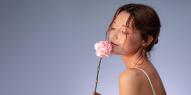 Parlak tenli, kapalı gözlü ve yüzüne nazikçe dokunan pembe çiçekli zarif bir kadının stüdyo görüntüsü. Kadınlığı, sakinliği ve romantizmi yakalar. Güzellik ve cilt bakımı için mükemmeldir..