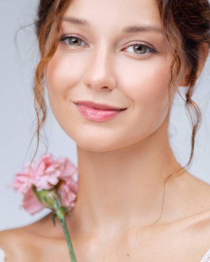 Güzellik ve sıcaklık saçan genç bir kadının zarif yakın plan portresi. Nazik gülüşü, parlak teni ve yumuşak göz makyajı yanında duran pembe karanfil çiçeğiyle tamamlanıyor.