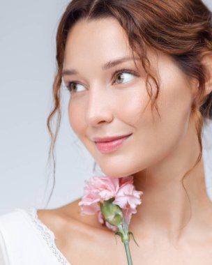 Çenesinin yanında pembe bir karanfil çiçeğini tutarken hafifçe gülümseyen genç bir kadının taze ve büyüleyici yakın çekimi. Işıl ışıl cildi, yumuşak makyajı ve gevşek ifadesi doğal bir güzellik yayıyor.