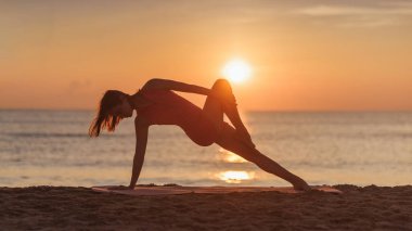 Pembe aktif giysili bir kadın silueti gün batımında plajda yoga pozu verir. Sakin okyanus ve parlayan gökyüzü barışçıl atmosfere eklenerek bu görüntünün yükseltilmesi için ideal hale geliyor.