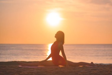 Yoga yapmış bir kadının silueti güneşin doğuşuyla plajda poz veriyor ve güneş vücudunu aydınlatıyor. Bu imaj, refahı, esnekliği, farkındalığı ve barışı teşvik etmek için idealdir.
