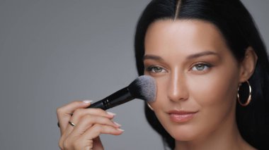 Sofistike bir kadın, kusursuz cildini vurgulamak için yumuşak bir fırçayla makyaj yapıyor. Zarif halka küpeler takıyor. Parlak bir teni var. Lüks güzelliği ve cilt konsepti var..