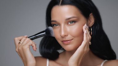 Göz kamaştırıcı genç bir kadının makyaj fırçasıyla pudra sürerken, kusursuz ve parlak cildini geliştirdiği yakın plan. Üst düzey kozmetik ve güzellik pazarlama kampanyaları için mükemmel.. 