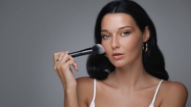 Kusursuz cildi olan zarif bir kadın yumuşak bir fırça kullanarak makyaj yapıyor. Altın halkalı küpeler ve beyaz bir bluz giyiyor. Lüks, güzellik eğilimleri ve profesyonel cilt kavramları var.. 