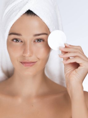 Kafasında beyaz bir havlu olan ve elinde pamuk pedi tutan beyaz tenli güzel beyaz bir kadın. Güzellik, cilt bakımı, yüz bakımı ve kozmetik reklamları için harika..