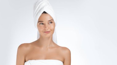 Beyaz bir havluya sarılmış, parlak ciltli genç bir kadın, hafif bir gülümsemeyle düşünceli bir şekilde ayakta duruyor. Saçları bir havluya sarılı, spa ve sağlık konsepti veriyor. Güzellik reklamları için mükemmel..