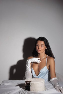 Mavi saten elbiseli ve uzun eldivenli güzel bir Asyalı kadın pastayla poz veriyor. Moda editörlüğü, lüks yaşam tarzı veya kutlama tanıtımları için mükemmel zarif ve şehvetli bir konsept..