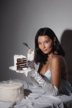 İpek mavisi elbisesi ve saten eldivenleriyle çekici esmer baştan çıkarıcı bir şekilde bir dilim çikolatalı pastayla poz veriyor. Yatak odası başyazıları, lüks moda veya doğum günü temalı görseller için ideal.