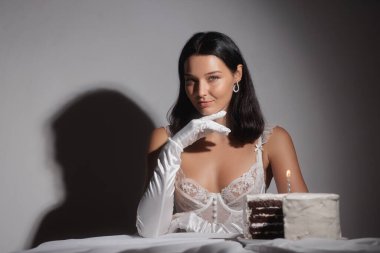 Beyaz dantelli iç çamaşırlı ve eldivenli, loş ışıkta mum ve pastayla poz veren baştan çıkarıcı genç kadın. Reklamlar, tanıtım ve editoryal kullanım için kavramsal doğum günü, lüks ve güzellik fotoğrafı.