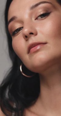 Işıl ışıl cildi, yumuşak altın göz makyajı ve büyük halka küpeleri olan kendine güvenen bir kadının yakın portresi. Güzellik, moda veya lüks marka tanıtımı için yüksek kaliteli stüdyo çekimi.