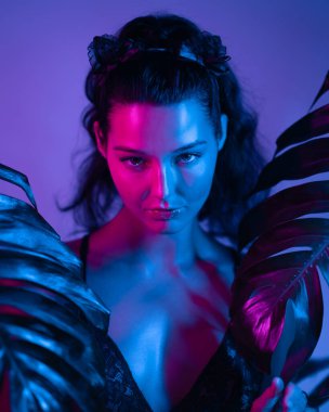 Neon mavisi ve pembe ışıkla aydınlatılan şehvetli model tropikal yapraklarla poz veriyor. Modern, canlı bir görünüm arayan moda, editör ve sanatsal projeler için idealdir..