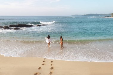 İki kadın, bikini giymiş, biri beyaz, biri kırmızı, sakin bir tropikal plajda okyanusa doğru yürüyor. Görüntü, yaz rahatlığını ve deniz kenarında olmanın huzurlu neşesini güzel bir şekilde tasvir ediyor..