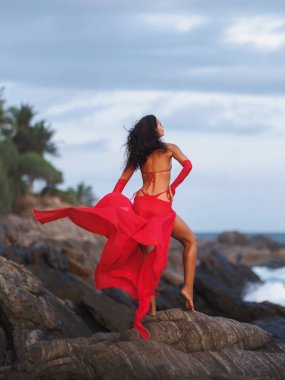 Kırmızı elbiseli ve bikinili çekici bir kadın kayalık sahilde poz veriyor. Yüksek kalite kavramsal moda fotoğrafı stil editörleri, seyahat reklamları veya duygusal yaşam tarzı promosyonları için idealdir..