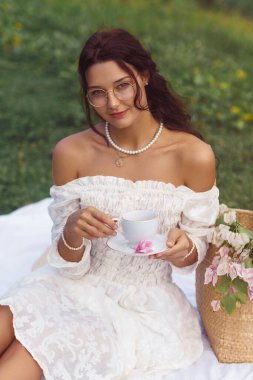 Beyaz, omuzsuz bir elbise içindeki zarif genç kadın piknik battaniyesinde çiçekli ve kamış çantalı çayın tadını çıkarıyor. Yaşam tarzı, zarafet veya romantik açık hava teması için ideal..