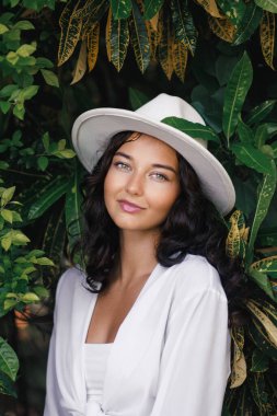 Beyaz şapkalı ve kıyafetli çarpıcı bir kadın gür tropikal yaprakların önünde zarifçe poz veriyor. Moda, güzellik ve yaşam tarzı kampanyaları için mükemmel. Doğal zarafeti, tarzı ve açık havayı vurguluyorlar.