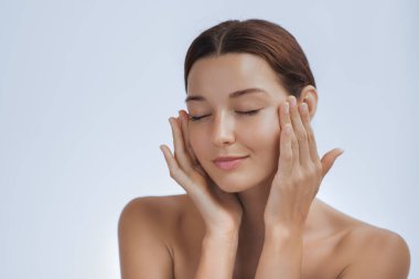 Gözleri kapalı, ışıldayan tenine nazikçe dokunan, rahatlatıcı ve sakin bir kadın. Güzellik, cilt bakımı ve hidrasyon ve yüz bakımına odaklanmış kişisel bakım reklamları için mükemmel..