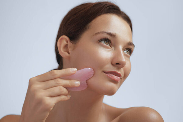 Young Caucasian woman massaging her face with a pink gua sha stone tool to enhance skincare, promote lymphatic drainage, and facial lifting. Ideal for spa promo and beauty product ads.