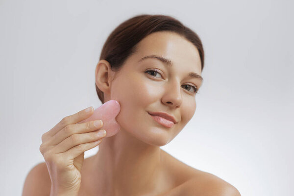 Young woman with glowing skin gently massages her cheek with a pink stone gua sha tool, promoting self-care, skincare routine, and natural beauty- ideal for spa, wellness, and cosmetic brands.