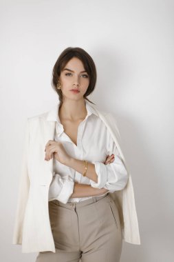 Beyaz bir ceket, beyaz gömlek ve bej pantolon giyen sofistike bir esmer kadın, kollarını minimalist beyaz bir arka plana çarpmış şekilde güven içinde poz veriyor. Moda ve yaşam tarzı temaları için mükemmel
