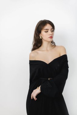  Güzel bir esmer kadın, siyah, omuzsuz bir elbise ve altın mücevherler giyiyor. Minimalist beyaz bir arka plana karşı zarafetle poz veriyor. Güzellik, moda ve sofistike temalar için mükemmel. 
