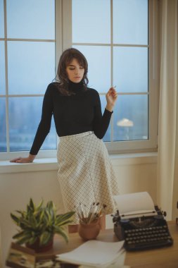 Siyah balıkçı yaka ve kareli etek giyen genç beyaz bir kadın klasik daktiloyla yaratıcı bir çalışma alanının penceresinde dikiliyor. Yaşam tarzı, yazı işleri ve yaratıcı kavramlar için mükemmel.. 