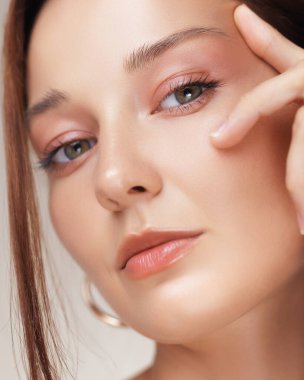 Şeftali rengi makyajlı, parlak tenli ve göz bölgesine dokunurken kaldırılmış bakışlı bir kadının yakın çekim stüdyosu portresi. Cilt bakımı, yaşlanmayı önleme ve kozmetik kampanyaları için ideal..