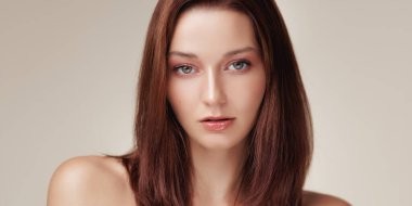 Parlak tenli, parlak kahverengi saçlı, kendine güvenen beyaz bir kadın temiz bir stüdyo ortamında kameraya ciddi ciddi bakıyor. Cilt bakımı, lüks güzellik reklamları ya da editoryal kullanım için mükemmel..