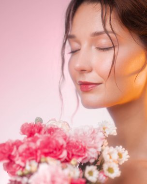 Güzel, genç, beyaz bir kadın rüya gibi, yumuşak odaklı stüdyo sahnesinde taze çiçeklerin kokusundan hoşlanıyor. Cilt bakımı, güzellik reklamları, spa tanıtımları ve romantik yaşam tarzı içeriği için ideal..