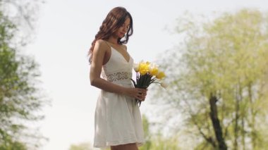 Beyaz elbiseli genç bir kadın güneşli bir parkta sarı ve beyaz laleler tutuyor. Bahar promosyonları, yaşam tarzı reklamları ve yüksek kaliteli doğa görselleri için ideal..