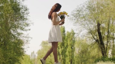 Beyaz elbiseli genç bir kadın güneşli bir parkta duruyor sarı ve beyaz lale buketini kokluyor. Bahar promosyonları, yaşam tarzı reklamları ve yüksek kaliteli doğa görselleri için ideal..