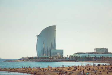 Barcelona 'daki güneşli plaj, kıyı şeridinin keyfini çıkaran insanlarla dolup taşarken, arka planda ikonik W Hotel kulesi yükseliyor. Seyahat reklamları, yaşam tarzı makaleleri ve yaz promosyonları için mükemmel..