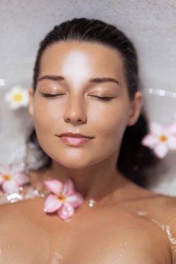 Çiçeklerle dolu bir banyoda yatan parlak tenli beyaz bir kadının yüksek kaliteli görüntüsü. Güzellik, spa, sağlık, özgüven ve doğal kozmetik temalar için mükemmel..