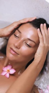 Güzel bir kadın rahatlatıcı bir banyodan zevk alıyor. Pembe tesisat çiçekleriyle dolu bir kaplıca küvetinde saçlarına hafifçe dokunuyor. Cilt bakımı ve lüks spa tanıtımları için yüksek kaliteli sağlık konsepti.
