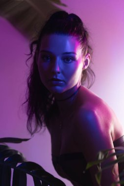 Tropikal yaprakların arasından canlı pembe ve mor neon ışıkla aydınlatılan bir kadının stilize edilmiş yakın çekimi. Moda yazıları, gece hayatı temaları ve egzotik tanıtım kampanyaları için harika..