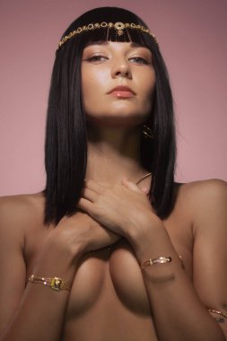 Parlak siyah saçlı ve altın takılarla süslenmiş güzel bir kadın pembe bir fona zarif bir şekilde poz veriyor. Onun çıplak omuzları ve dengeli ifadesi lüks ve sofistike bir duygu iletir. İdeal