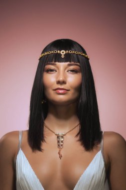 Pembe arka planda, zarif siyah saçları, zarif makyajı ve karmaşık altın takıları olan Kleopatra gibi bir kadın poz veriyor. Bu resim zamansız güzelliği ve görkemli sofistike görüntüyü yakalar.