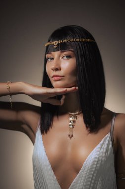 Pürüzsüz siyah saçlı, Kleopatra 'dan esinlenilmiş altın başlık ve kolyeyle süslenmiş güzel bir kadın zarif bir poz veriyor, elleriyle yüzünü çerçeveliyor. Lüks tarzı ve özgüveni