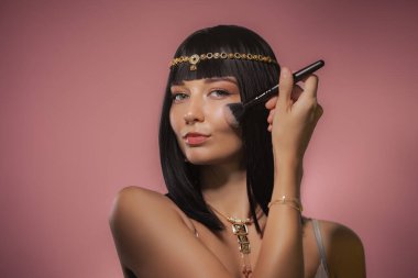 Kleopatra 'dan esinlenilmiş altın başlık ve kolyeyle süslenmiş, zarif, siyah saçlı bir kadın pembe arka planda bir fırçayla makyaj yapıyor. Kendine güveni ve şatafatı