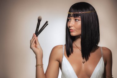 Parlak siyah saçlı, Kleopatra 'dan esinlenilmiş altın başlık takan güzel bir kadın pembe arka planda kendine güvenen bir makyaj fırçası tutuyor. Özgüvenli ifadesi ve lüks tarzı.